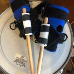 a pair of small item aids holding two drumsticks resting on a drum - don't let reduced hand function stop you from drumming!