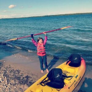 Child on the shore next to a yellow kayak. She is holding her paddle above her head and grinning. She uses a Limb Difference gripping aid in her right hand.