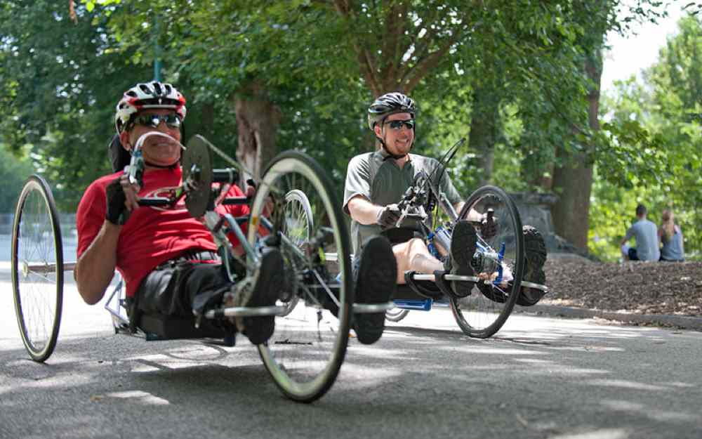 two smiling men riding their hand bikes down a tree-lined path