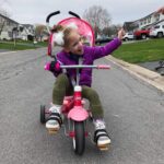 Image shows a girl riding a trike and waving, joyfully. She is gripping the right handlebar with a pink Active Hands mini gripping aid.