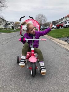 Image shows a girl riding a trike and waving, joyfully. She is gripping the right handlebar with a pink Active Hands mini gripping aid.