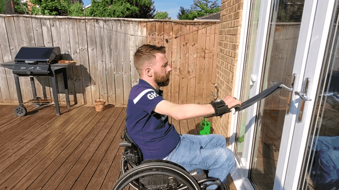 A gif showing Gareth doing a one arm row outside on his decking. He uses the door hand to hold one end of a theraband and a hook aid for the other