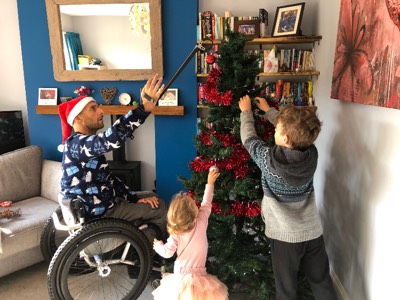 Image shows man in wheelchair wearing Christmas hat, using a reacher to decorate a christmas tree. His two children are helping.