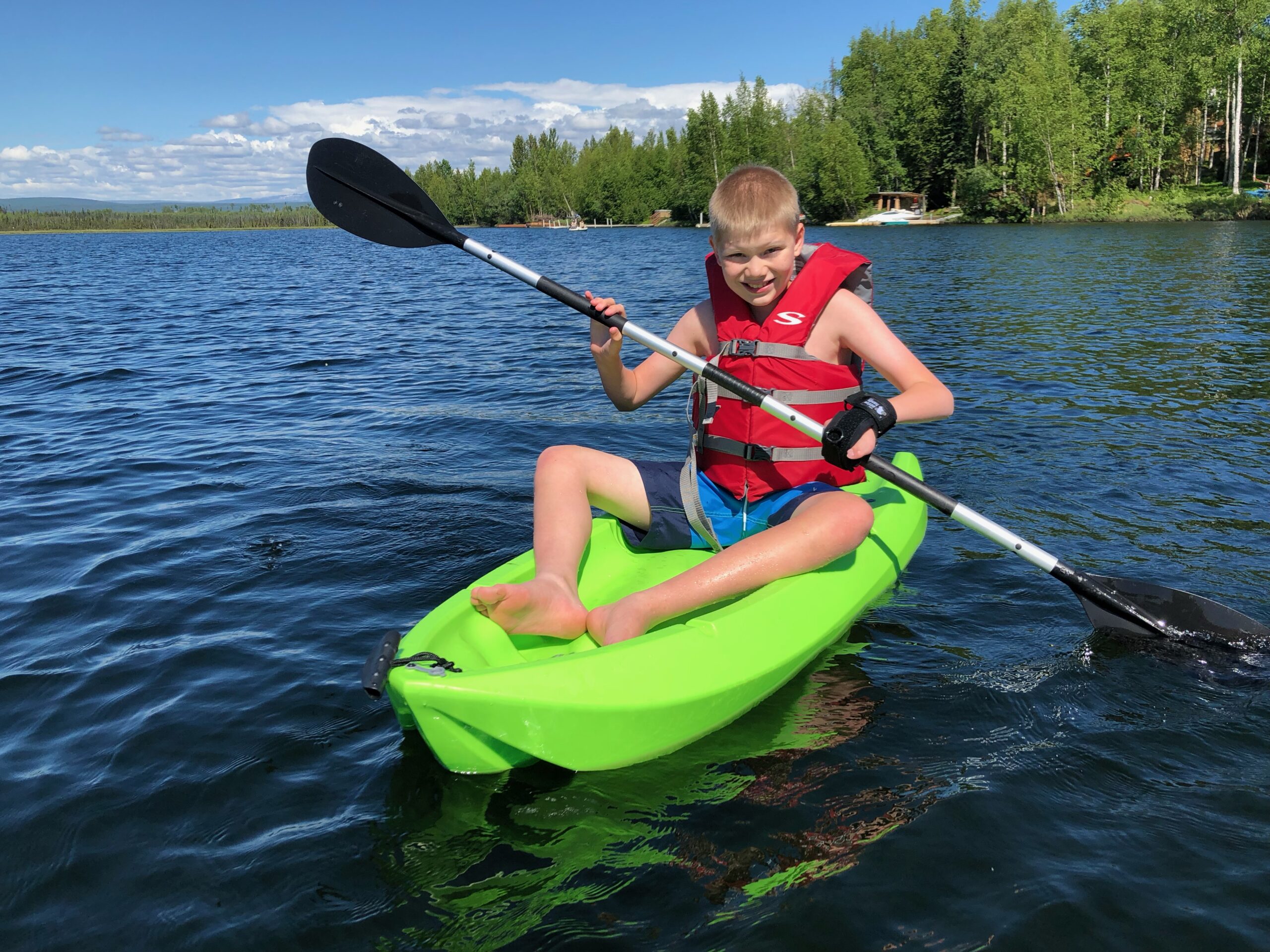 Boy sits in a bright green kayak holding the paddle with a limb difference gripping aid on his left hand.