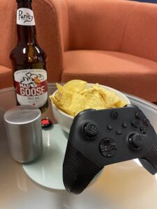 Playstation controller with gaming grips attached, bowl of nachos, open bottle of beer and automatic bottle opener are all arranged on a glass coffee table.