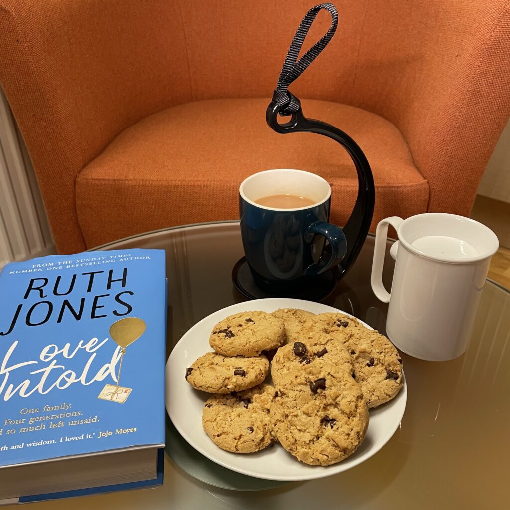 A self-care scene of a comfy chair, book, cookies, a cup of tea on the spillnot and a hand steady mug