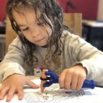 Child uses functional hand product to grip a crayon and colour in a picture.