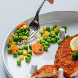Image shows chicken and vegetables in the One-Handed Plate. A fork uses the high sides to scoop peas, while the chicken is held in place by the pegs, ready for cutting one-handed.