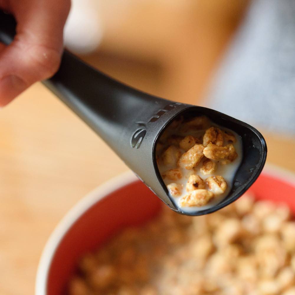 A black spoon with a cavity inside it holds a scoop of cereal and milk with the bowl in the background. The cavity inside the spoon ensures the food stays inside the spoon and doesn't spill.