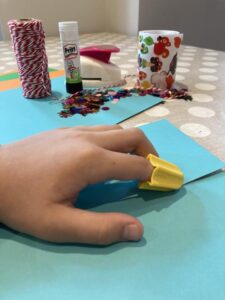 Crafting materials are laid out on a table and a child's hand is pictured using the Nimble one-finger cutter to cut the paper.