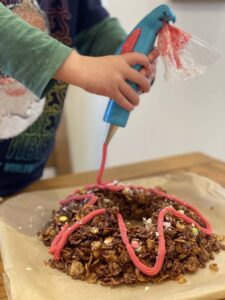 A child uses the icing pen to squeeze pink icing onto a cake below.