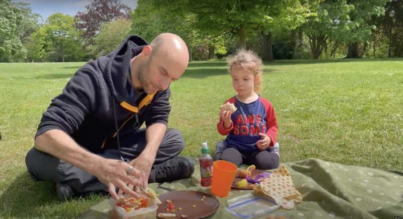 A dad and his young daughter are sharing a picnic. The dad uses cutlery with loops to overcome his reduced hand function, while serving pasta salad.