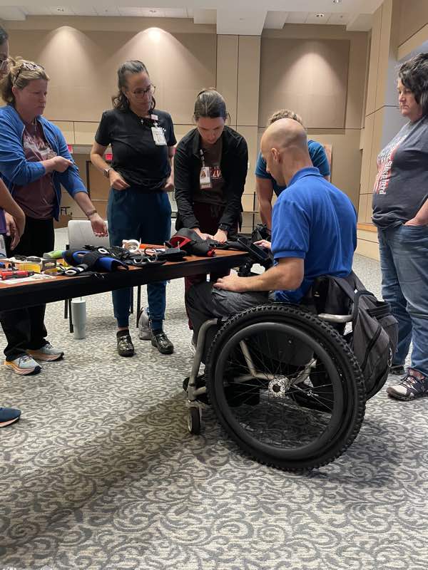 Rob sits at a table displaying a range of Active Hands products, while a team of OTs look in with interest.