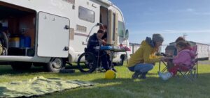 A motorhome is parked on a grass pitch. Rob is cooking breakfast at an outdoor stove and Jo is crouched in front of a toddler on a camping chair, passing her a plate of food. They are all wearing warm hats and coats, but the sun is shining.