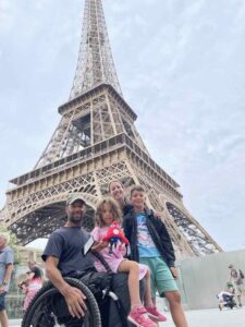 Rob, a wheelchair user, sits in front of the Eiffel Tower. His daughter is sat on his lap, his son and wife stand behind him.