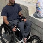 Rob, a wheelchair user, is pushing down a coastal path, next to a harbour wall. He has his phone in front of him, attached the Flexi Phone Holder, so he can navigate.