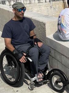 Rob, a wheelchair user, is pushing down a coastal path, next to a harbour wall. He has his phone in front of him, attached the Flexi Phone Holder, so he can navigate.