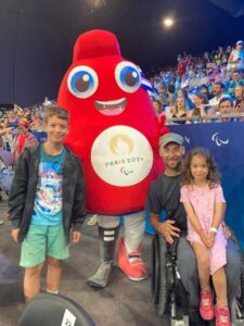 Rob, a wheelchair user, poses with a costumed Paralympic mascot. His daughter sits on his lap and his son stands the other side of the mascot. They are all smiling.