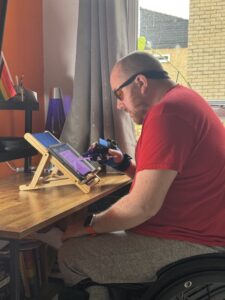 Oliver sits at a desk easel, using the small item aid to hold an Apple Pencil while completing a piece of art on his iPad.