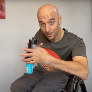 Rob, a wheelchair user, is looking directly at camera, spraying a deodorant, using an easy spray, at the camera.