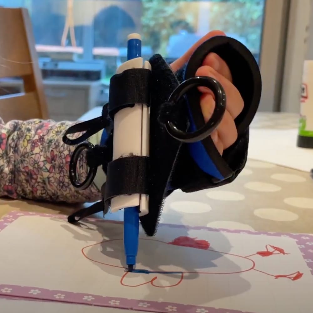 A blue coloring pen is being held in a clamp that is part of a gripping aid, worn by a child who is coloring a picture of a butterfly. The image is a close up of just the hand, gripping aid and picture.