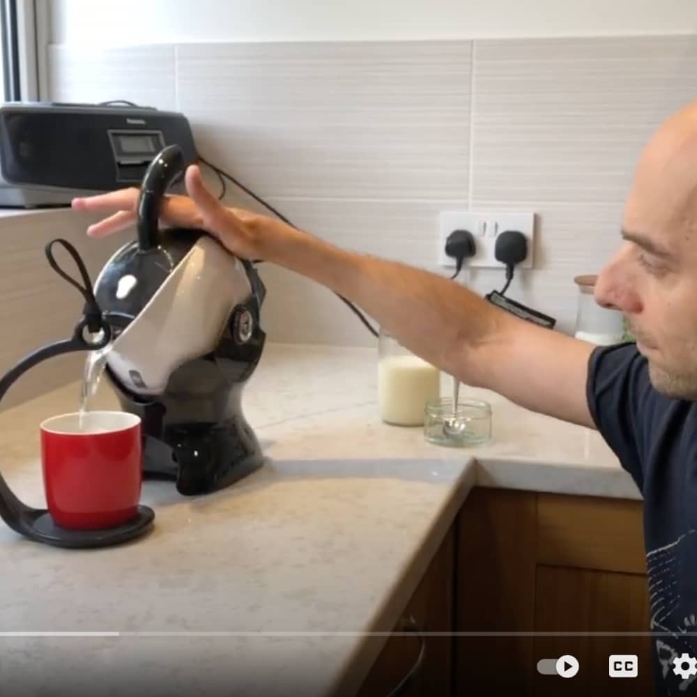 A man with reduced hand function pours water from a kettle into a mug by tipping the kettle in its special base. There is no need for him to lift the heavy kettle.