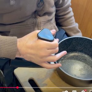 Image shows a hand holding a saucepan handle which has been fitted with the pan buddy - a device which fits over a regular saucepan handle to create a vertical handle with arm cuff support. Much easier to hold for those with reduced hand function.