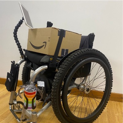 A manual wheelchair has a box on the seat and lots of great accessories attached to it, such as a phone mount, a cup holder and a lap strap to hold the box in place.