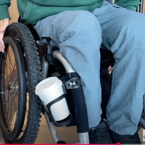 Image shows a manual wheelchair with someone sitting in it. One the frame at the front is a wheelchair drinks holder with a takeaway coffee cup in it.