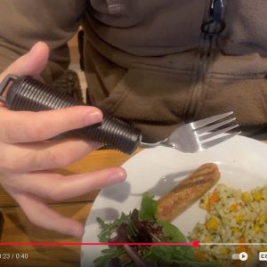 Image shows a fork with a chunky handle held above a plate of food. The fork has been bent towards the man's mouth. This means he doesn't need to bend his wrist to eat.