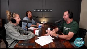 A man is interviewing two women for a podcast. They are sat around a desk, each with a microphone. They are smiling.