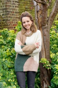 A woman stands in a leafy garden, arms folded gently across her chest, smiling at the camera.