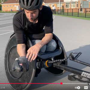 Rob is in a racing wheelchair and showing his bespoke-made racing gloves to the camera.