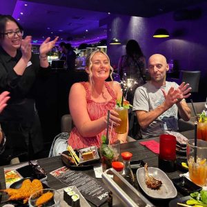 image shows millie and rob at a sushi restaurant, smiling, with a waitress behind them clapping. Millie is holding a birthday cocktail.