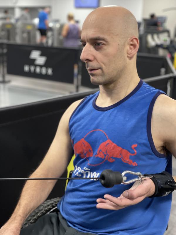 rob pulls the cable of a pulley machine towards his chest. He holds the cable using a D-ring aid