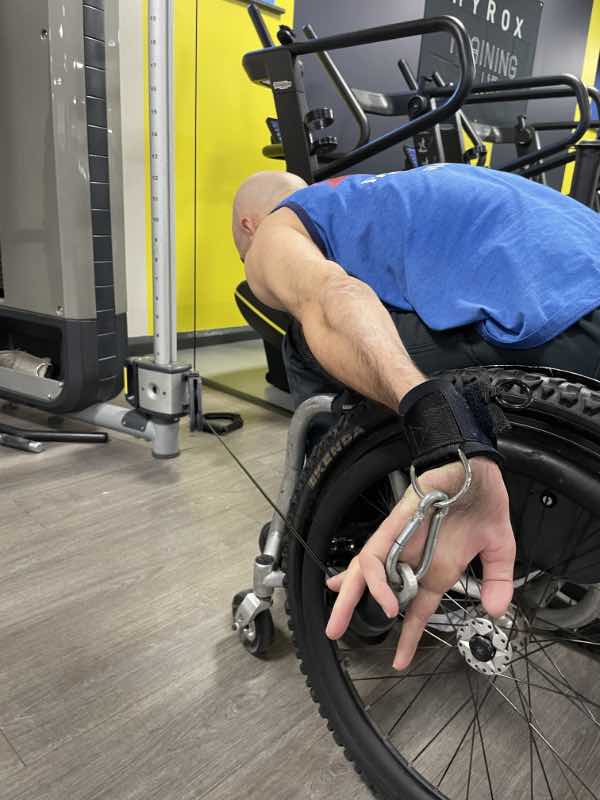 photo from behind rob, a wheelchair user, who has extended his arm behind him, holding the cable pf a pulley machine using a D-Ring aid.