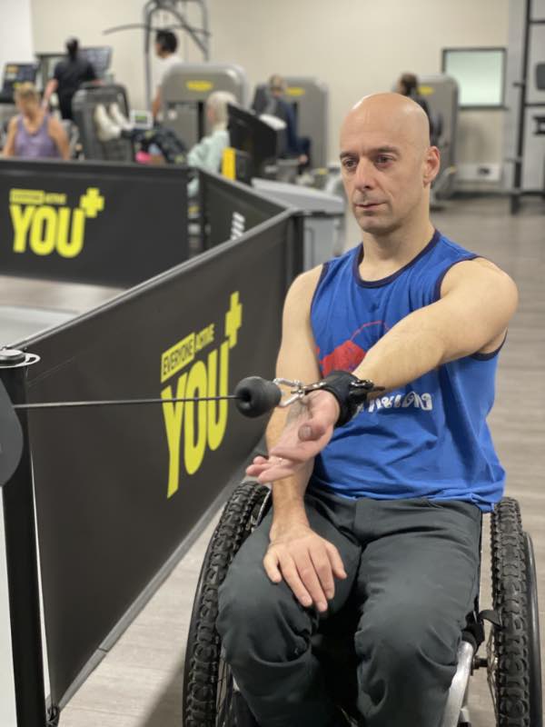 rob extends his arm in front of him, holding the cable of a pulley machine using a d-ring aid.