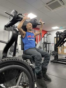 image shows rob working out doing an overhead press. His wheelchair waits beside the machine