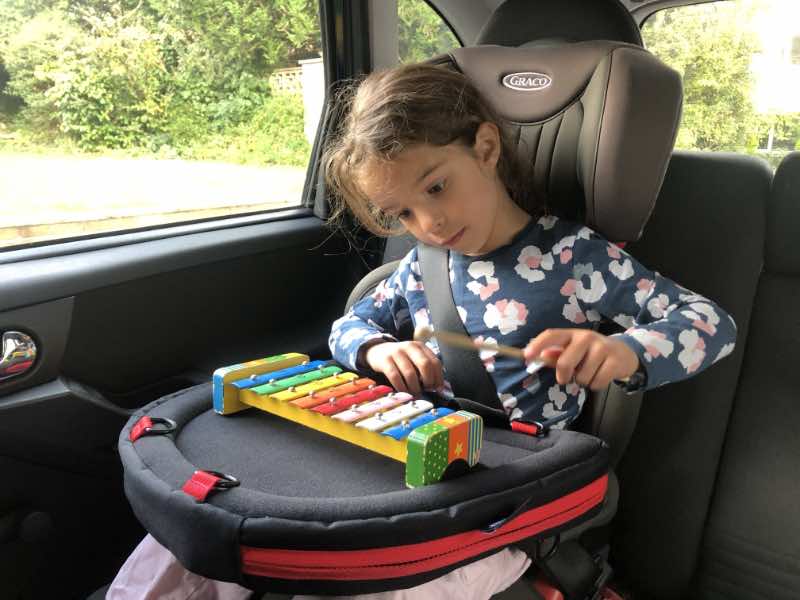 image shows a young child in a car seat with a Trabasack bag on her lap. The bag offers a surface to play her xylophone on.