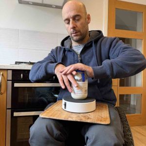 rob, a wheelchair user, has a wooden board on his lap. On this is the hold fast platform with a jar of mayonnaise on top. The hold fast is holding the jar while rob twists the lid off.