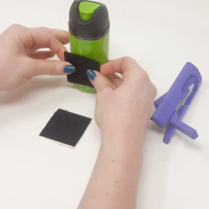 Image shows two hands putting adhesive Velcro onto a green spray canister. An EasySpray lies next to them, ready to put on the can.